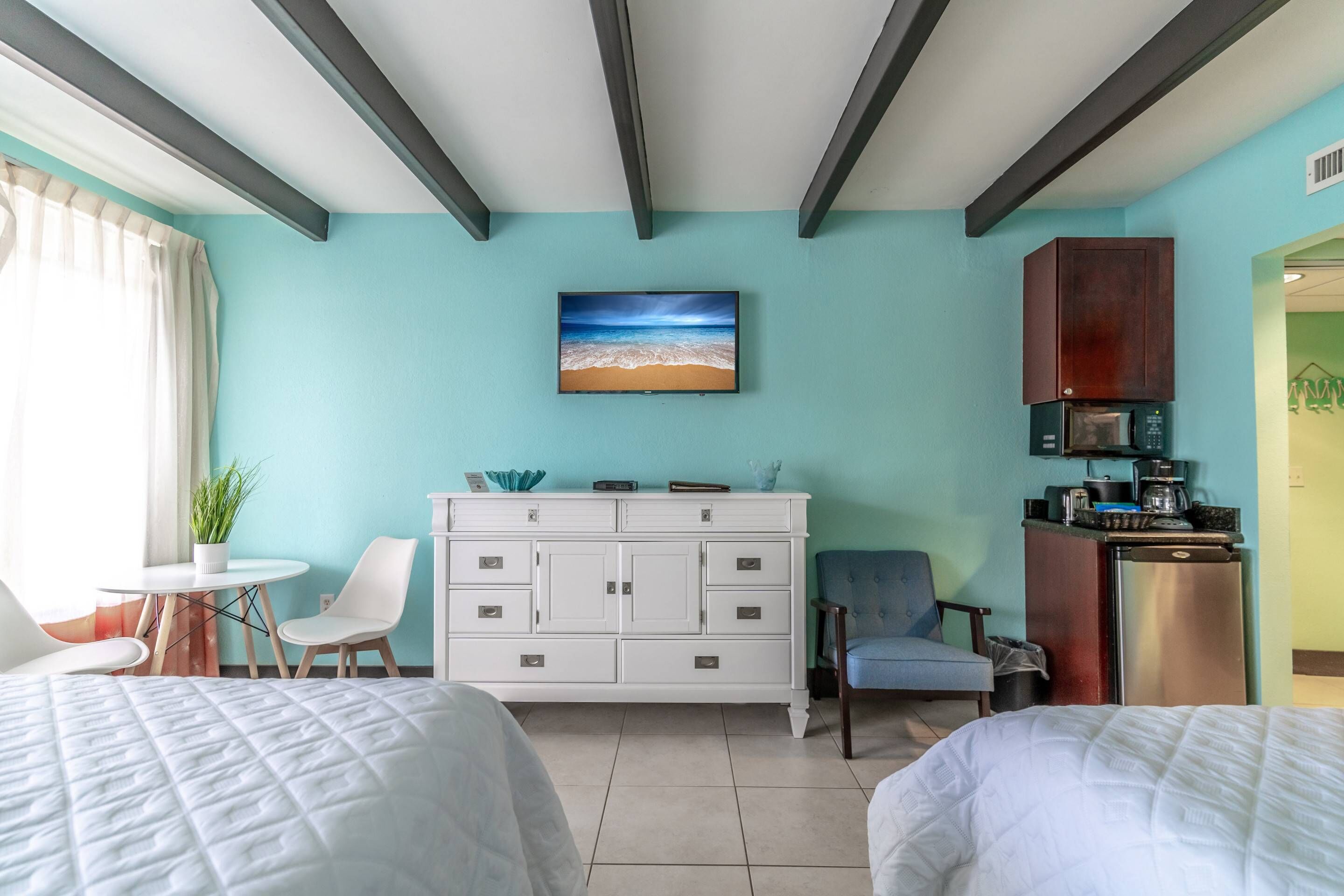 A hotel room with two beds, a white dresser, a small table with a plant, a blue chair, a TV on the wall, and a kitchenette with a microwave and coffee maker.