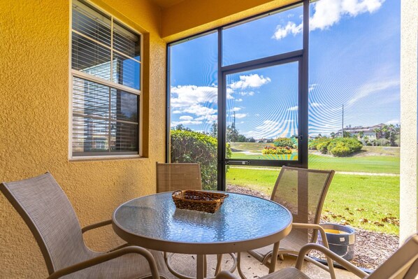 A covered patio with a round glass table, wicker chairs, and a view of a golf course through large windows.