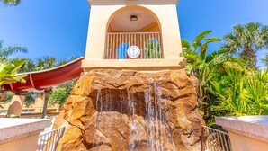 A water feature with a clock above it, surrounded by tropical plants.