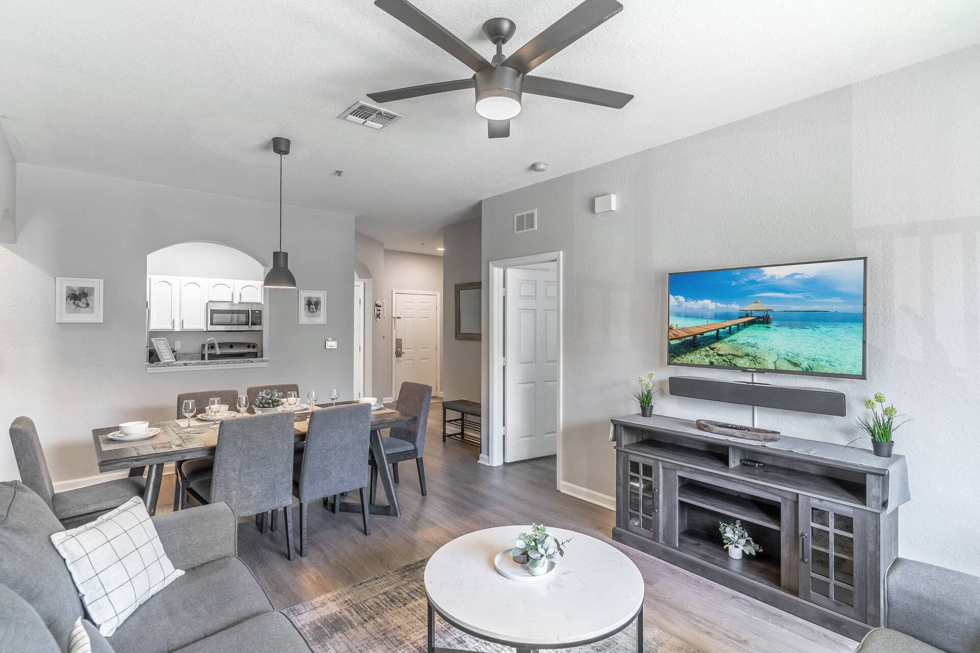 A modern living room with a flat-screen TV, a ceiling fan, and a dining area with a kitchen in the background.