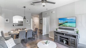 A modern living room with a flat-screen TV, a ceiling fan, and a dining area with a kitchen in the background. - 2821 Windsor Hills (Kissimmee)