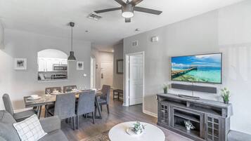 A modern living room with a flat-screen TV, a ceiling fan, and a dining area with a kitchen in the background.