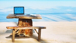 Ein Holztisch mit einem Laptop und Papieren, der auf einem sandigen Strand mit Blick auf den Ozean steht.