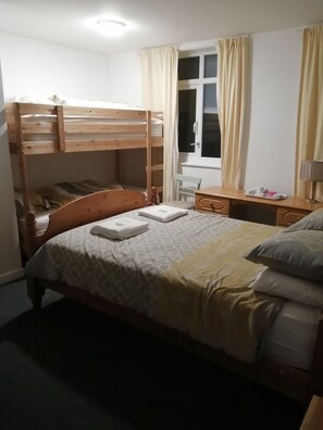 Una cama litera con cabecera de madera, una mesita de noche con lámpara y una ventana con cortinas.