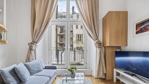 Ein modernes Wohnzimmer mit einer Couch, einem Couchtisch mit Pflanze, einem Fernseher auf einem Schrank und einem Fenster mit Vorhängen.