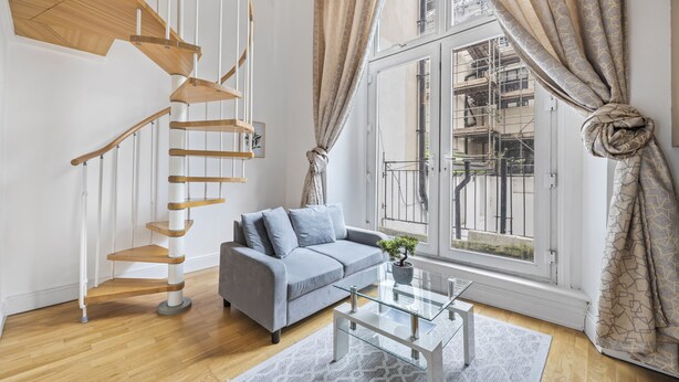 Ein modernes Wohnzimmer mit grauem Sofa, einem Glastisch, einer Holzspiralkonstruktion als Treppe und großen Fenstern mit Vorhängen.