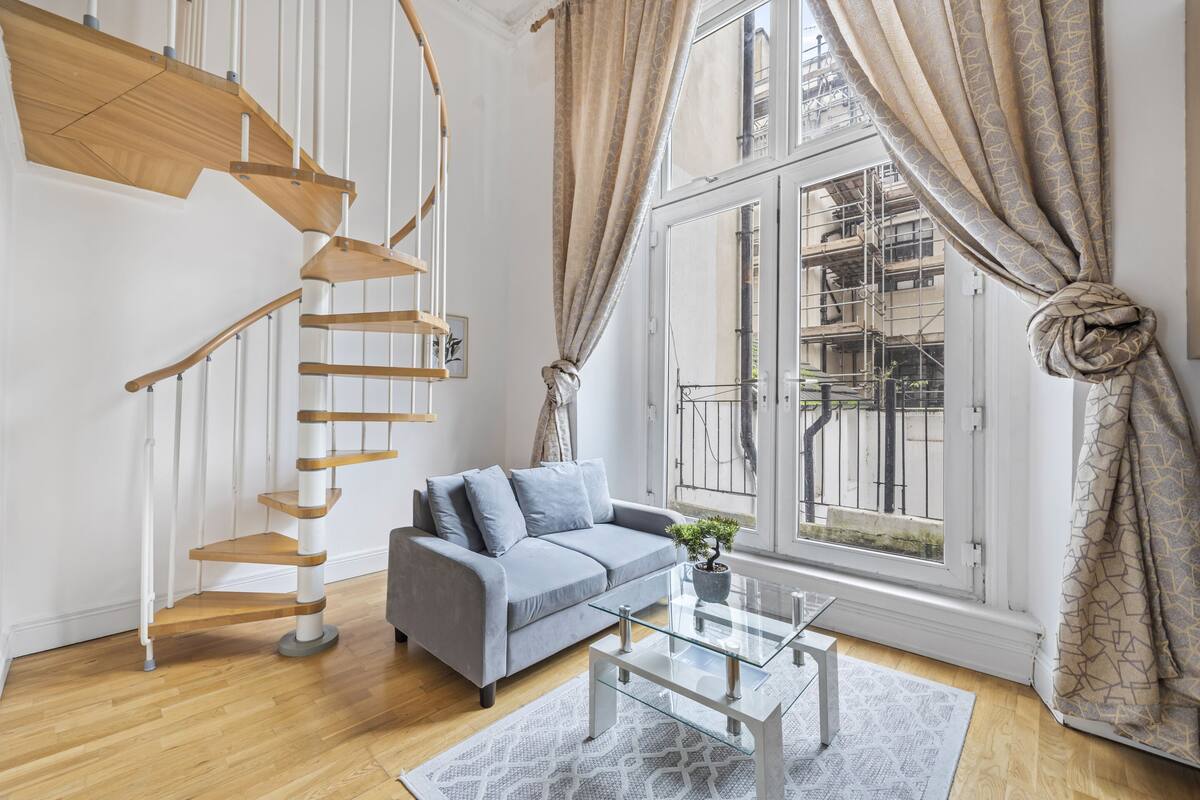 Un salon moderne avec un canapé gris, une table basse en verre, un escalier en colimaçon en bois et de grandes fenêtres avec des rideaux.