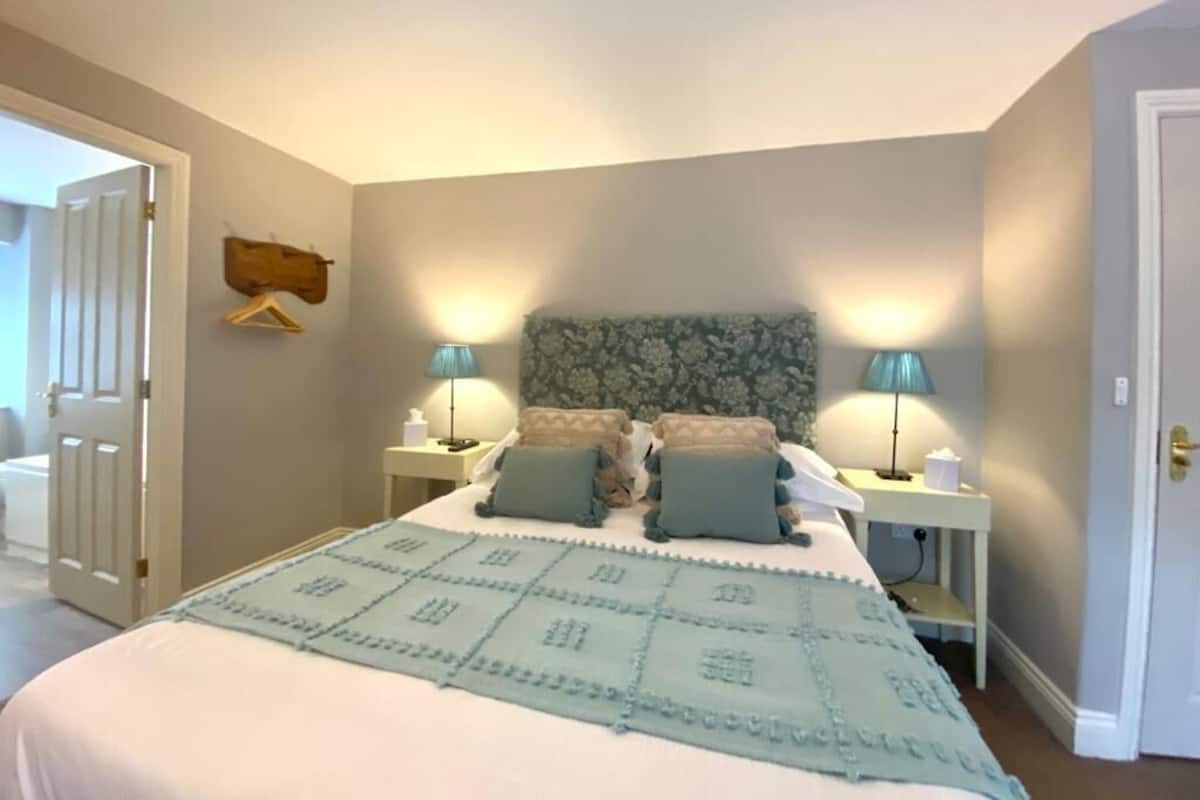 A bedroom with a bed, two bedside tables, a nightstand, a hanging coat rack, and a door.