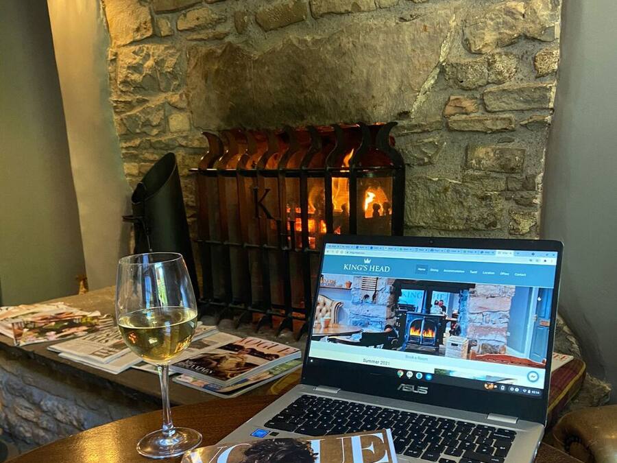A laptop on a table with a glass of wine, a magazine, and a stone wall with a fireplace in the background.