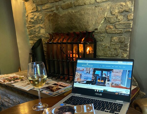 A laptop on a table with a glass of wine, a magazine, and a stone wall with a fireplace in the background. - The King's Head (Kirkby Stephen)