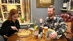 Una mesa de madera con pizza, vino y una vela. Dos personas están sentadas a la mesa.
