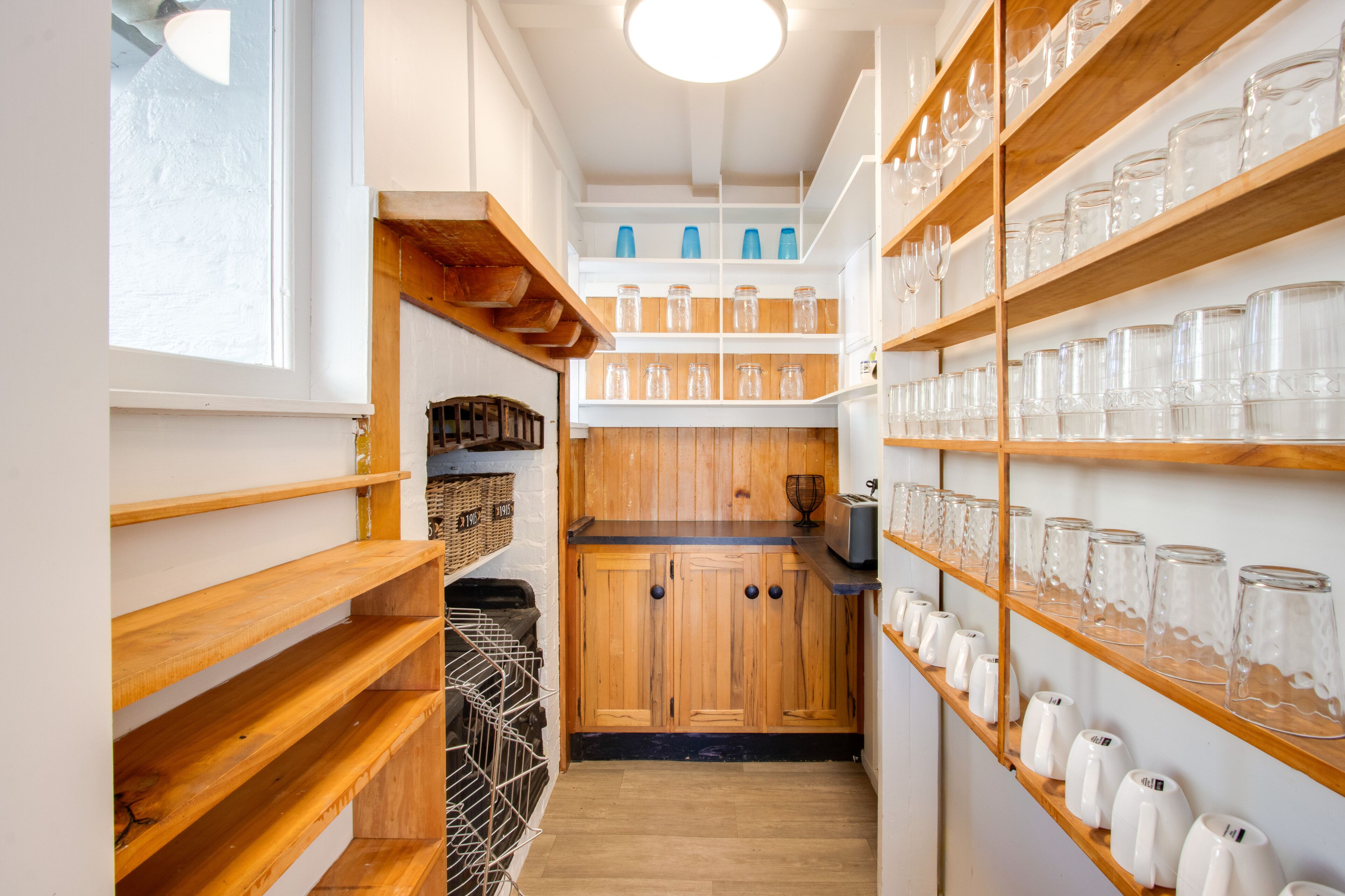 A narrow kitchen with wooden shelves and cabinets, glass jars, and a window.