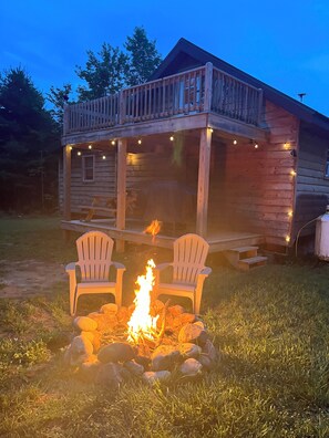 Una cabaña de madera con porche, dos sillas Adirondack y un brasero.
