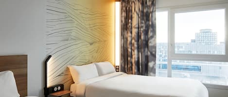 Una habitación de hotel con dos camas, una mesita de noche con lámpara, una obra de arte en la pared y un ventanal con vista a la ciudad.