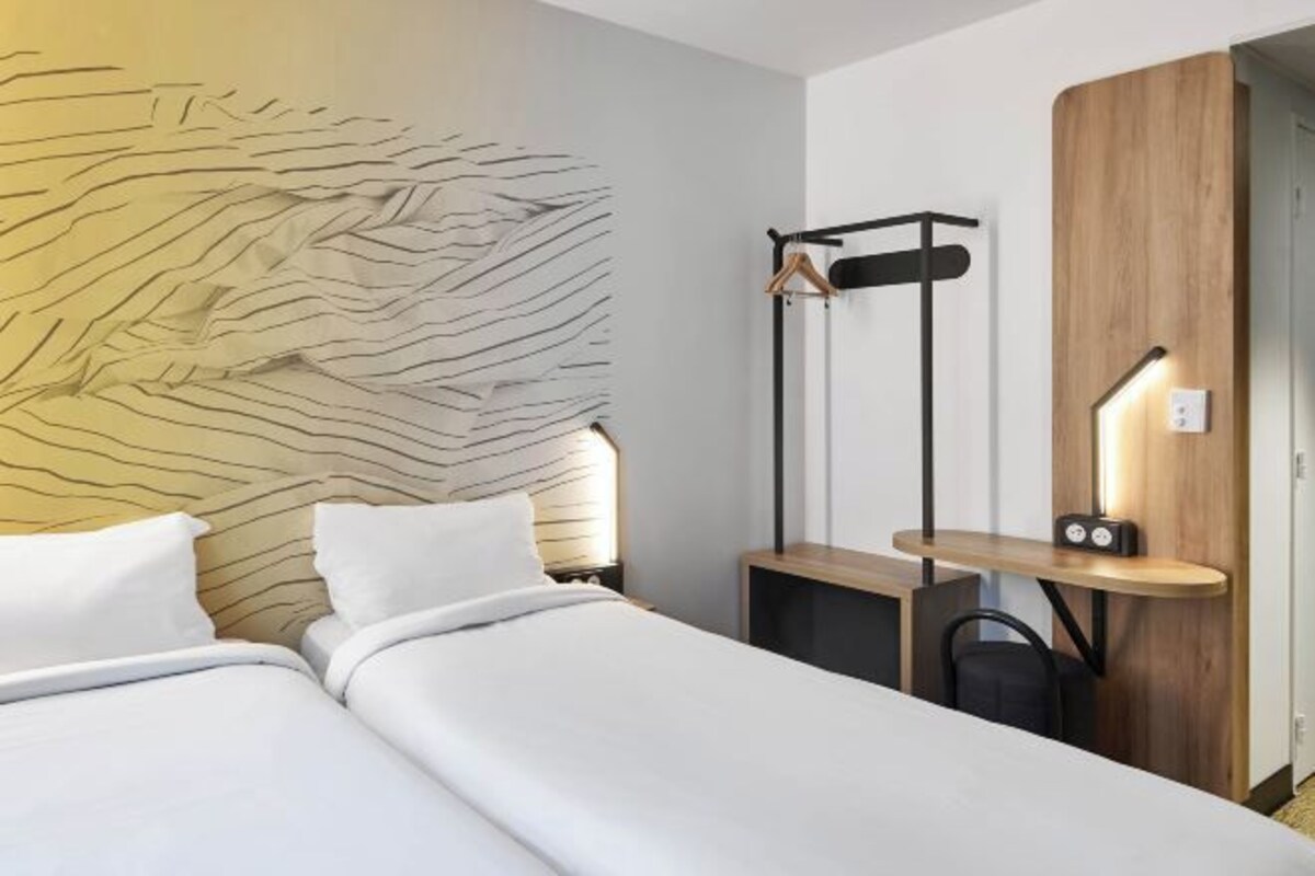 Una habitación de hotel moderna con una cama grande, un cabecero de madera, una mesita de noche con lámpara y un perchero.