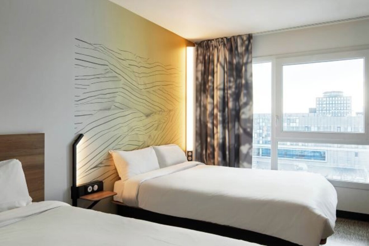 Habitación de hotel con dos camas, un cabecero moderno, un ventanal con vista a la ciudad y una pared con diseño texturizado.