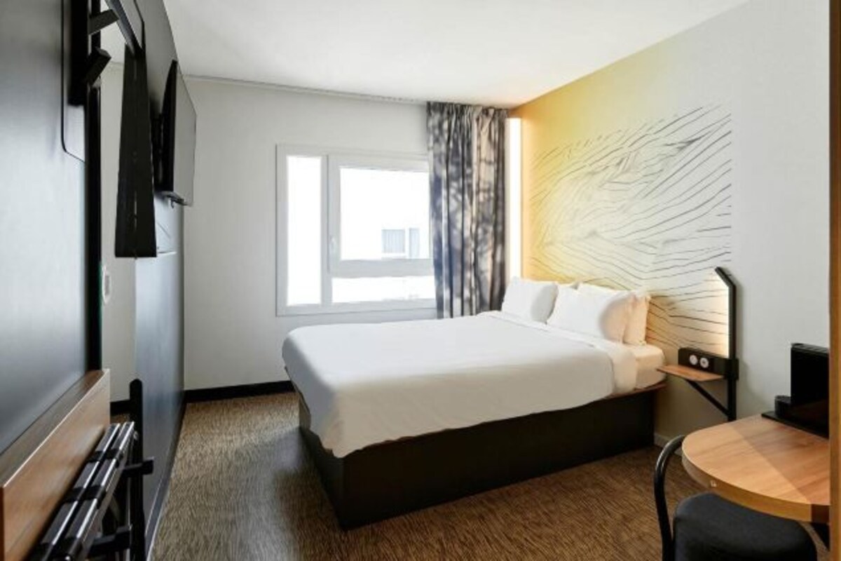 Habitación de hotel con cama, escritorio, silla, ventana y una obra de arte en la pared.