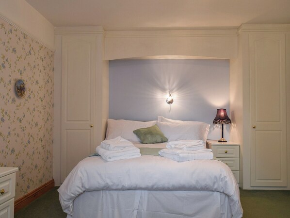 Ein ordentlich bezogenes Bett mit gefalteten Handtüchern, einer Nachttischlampe und einer Wandleuchte in einem Raum mit floralem Wandpapier und weißen Wänden.