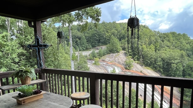 A wooden deck with a rustic table and chairs, overlooking a forested valley with a waterfall and a rocky outcrop.