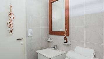 Um banheiro compacto com pia branca, espelho e vaso sanitário. Há uma pequena prateleira com um frasco e difusor de varetas. Um arranjo decorativo de conchas está pendurado na parede.