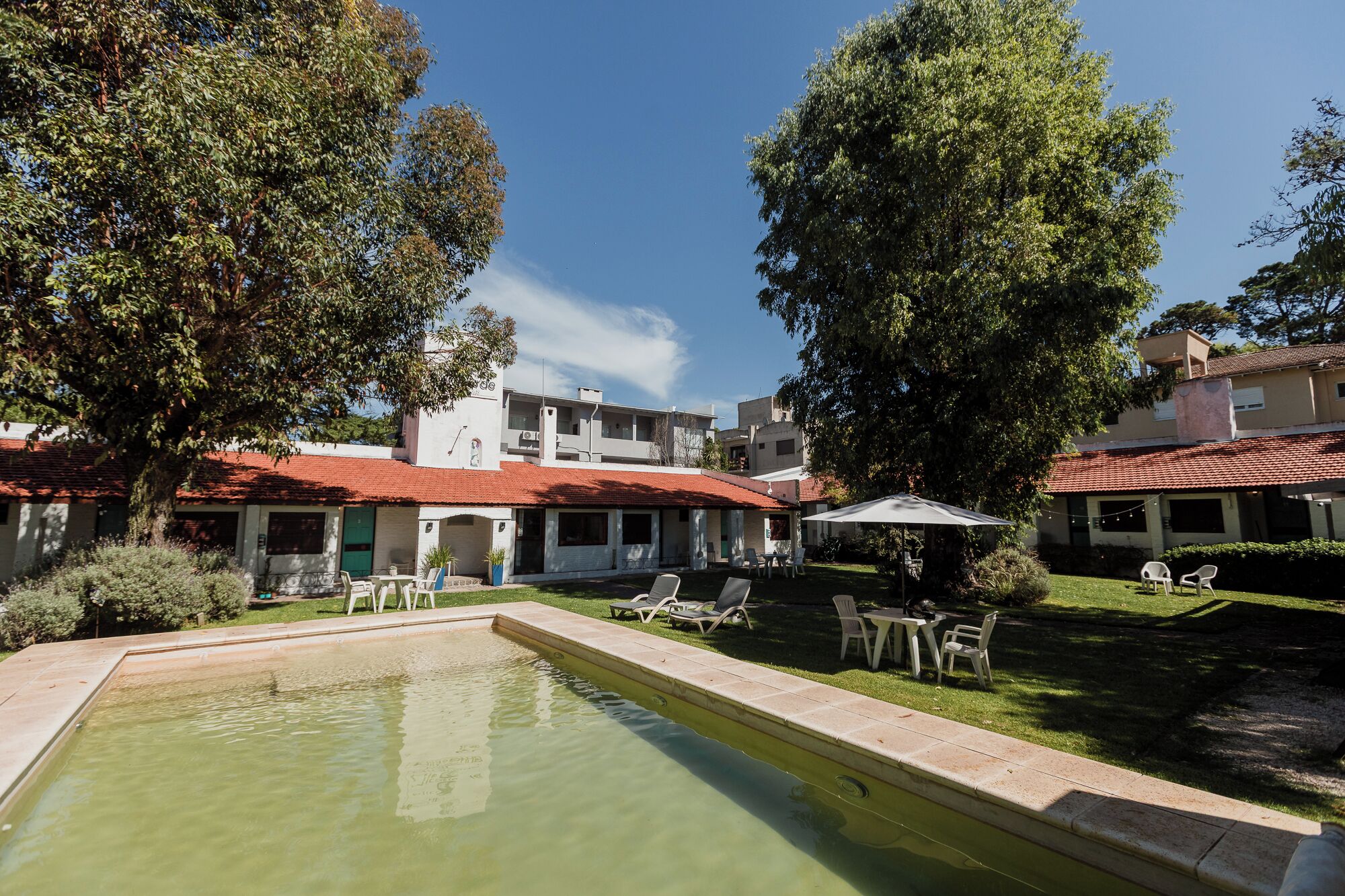 A pool area with lounge chairs and a small table, surrounded by greenery and buildings with red-tiled roofs.