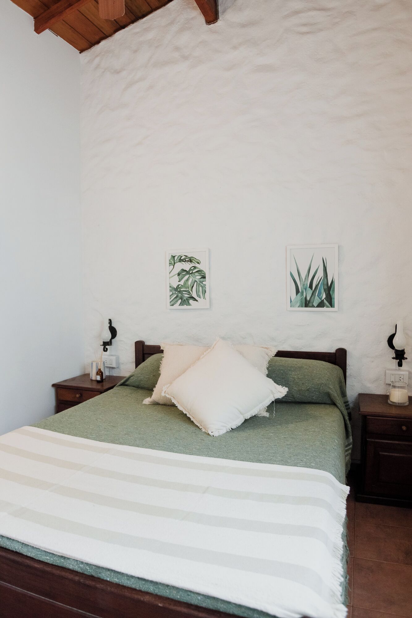 A bed with a green bedspread and white pillows, flanked by two nightstands with lamps and a small vase. Two framed botanical prints adorn the wall above the bed.
