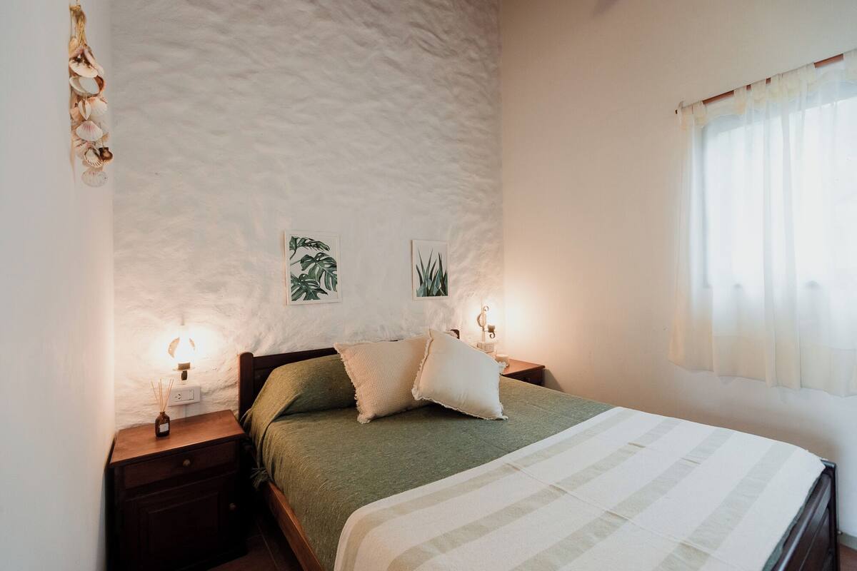 A bedroom with a bed, two wall-mounted nightstands, a window with sheer curtains, and two framed botanical prints on the wall.