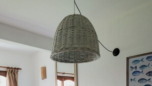 A room with a bed, a dining table with a placemat, a wicker hanging light fixture, a framed fish painting, and a window with curtains. - Miraverde Pinamar (Pinamar)