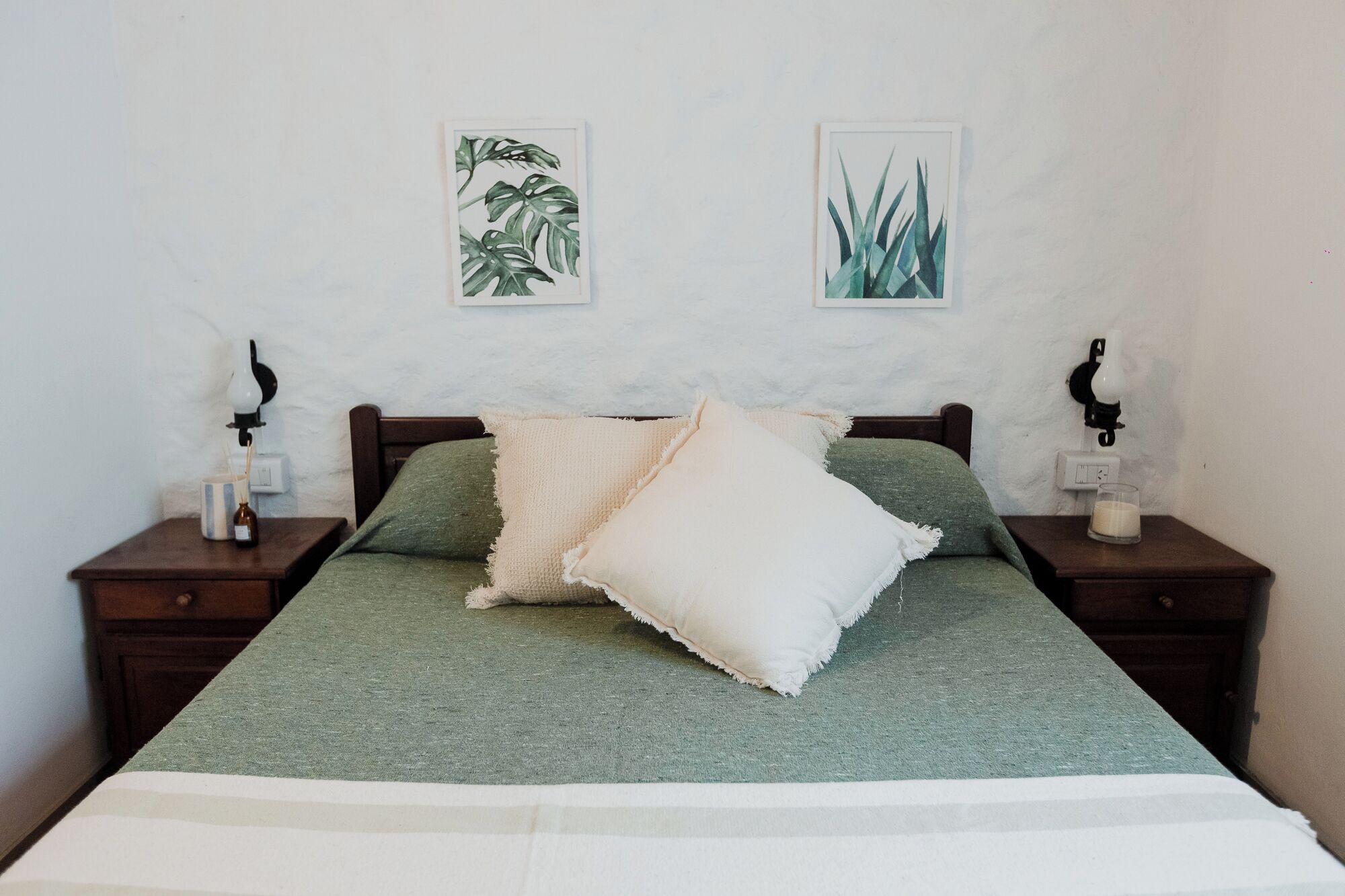 Una cama con una colcha verde y dos almohadas, flanqueada por mesitas de noche con lámparas de pared y una botella pequeña en cada una.