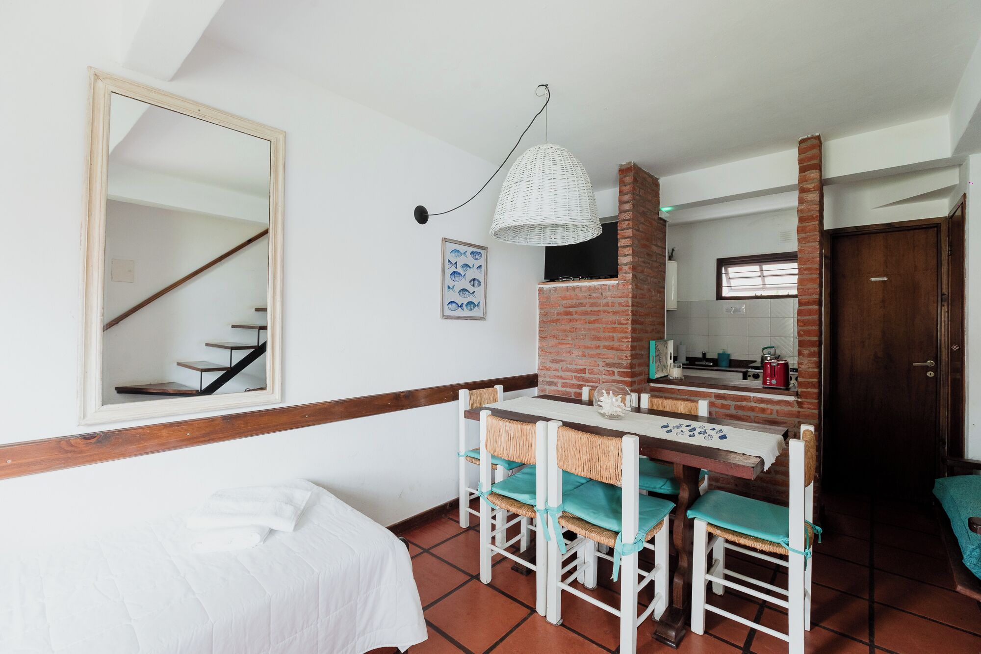A compact room with a bed, a dining table with chairs, a hanging light fixture, a brick fireplace, and a kitchen area visible through an open door.