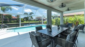 A poolside area with a view of a canal and houses, featuring a table and chairs, and a ceiling fan.