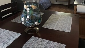 Una sala de estar con una mesa de comedor, un jarrón grande con piedras decorativas, un ventilador de techo, una ventana con cortinas, un radiador y una cama con una manta azul.