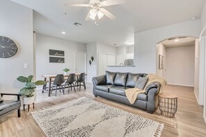 A modern living room with a grey leather sofa, a dining table with chairs, a large clock, and a ceiling fan.
