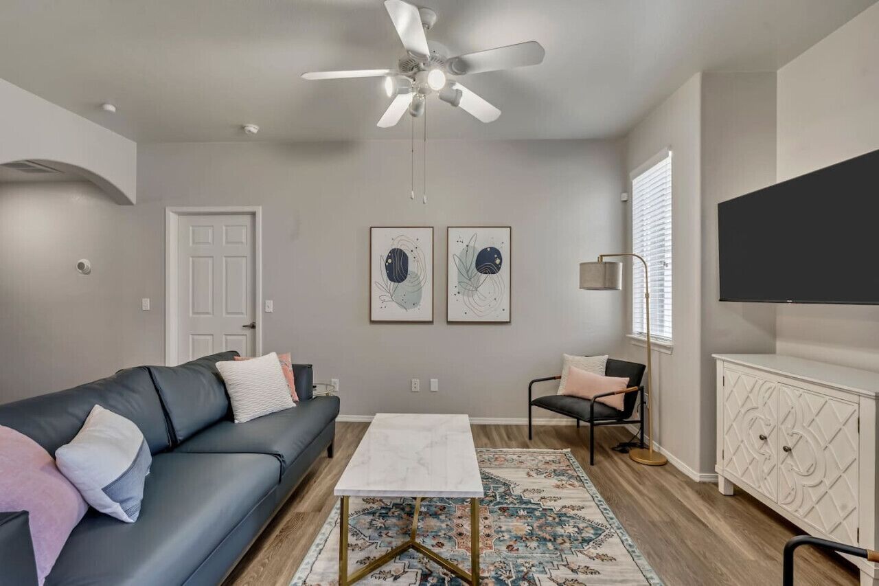 Un salon moderne avec un canapé gris, une table basse blanche, un tapis à motifs, un ventilateur de plafond et un téléviseur posé sur un meuble blanc.