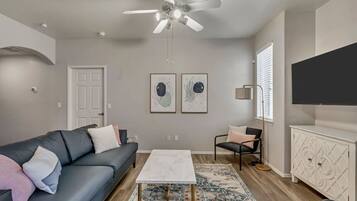 Un salon moderne avec un canapé gris, une table basse blanche, un tapis à motifs, un ventilateur de plafond et un téléviseur posé sur un meuble blanc.