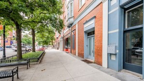 A sidewalk lined with trees and benches, adjacent to a row of brick buildings with large windows and a blue facade.