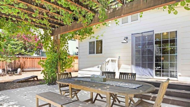 Eine Außenpatio mit Holztisch und -stühlen, eine Pergola mit Kletterpflanzen und ein weißes Gebäude mit Fenstern im Hintergrund.