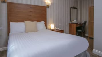 A hotel room with a large bed, a wooden headboard, a desk with a chair, a mirror, and a nightstand with a lamp.