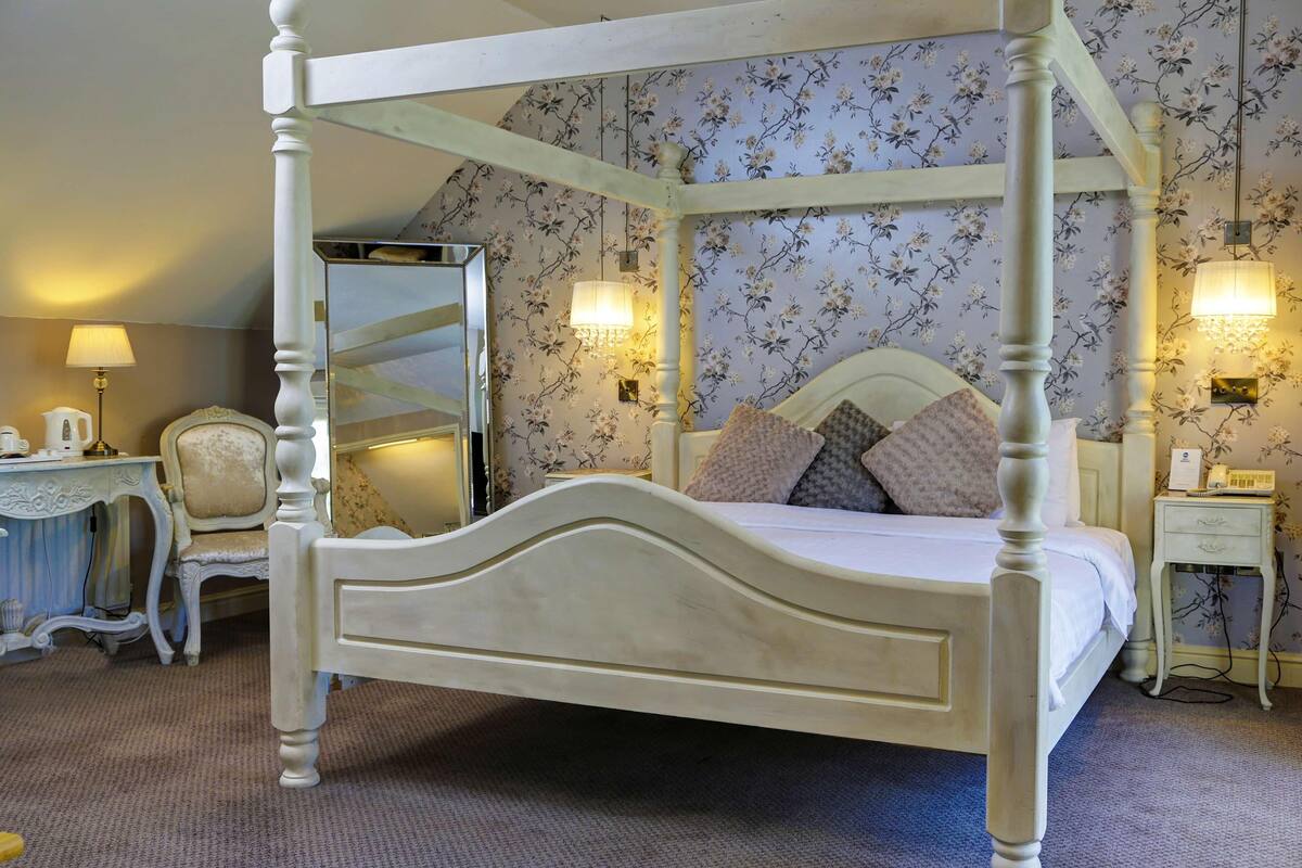 A four-poster bed with a mirror, floral wallpaper, bedside lamps, and a small bedside table with a telephone.