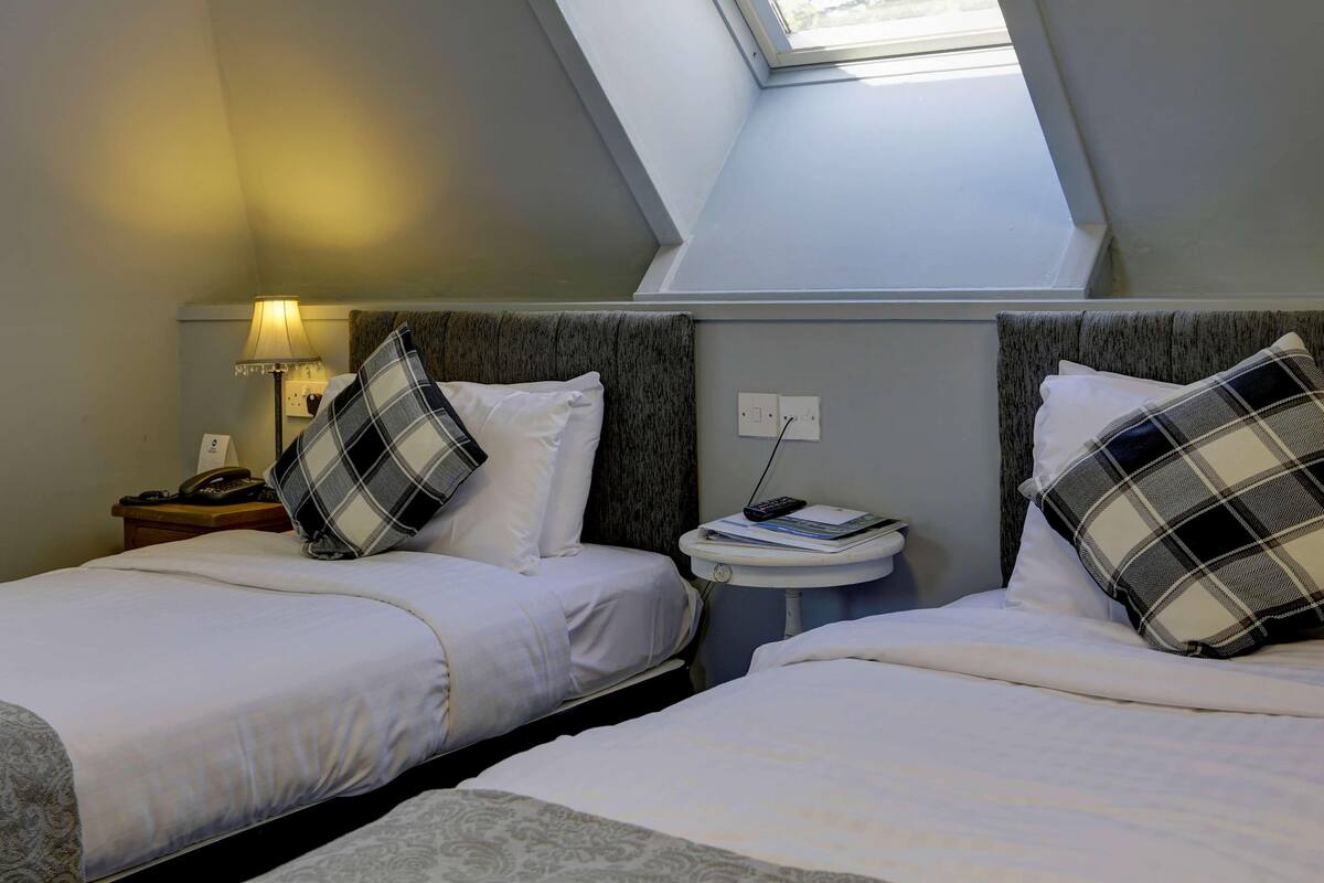 A hotel room with two beds, a nightstand with a lamp, a small table with a phone and a remote, and a skylight above.