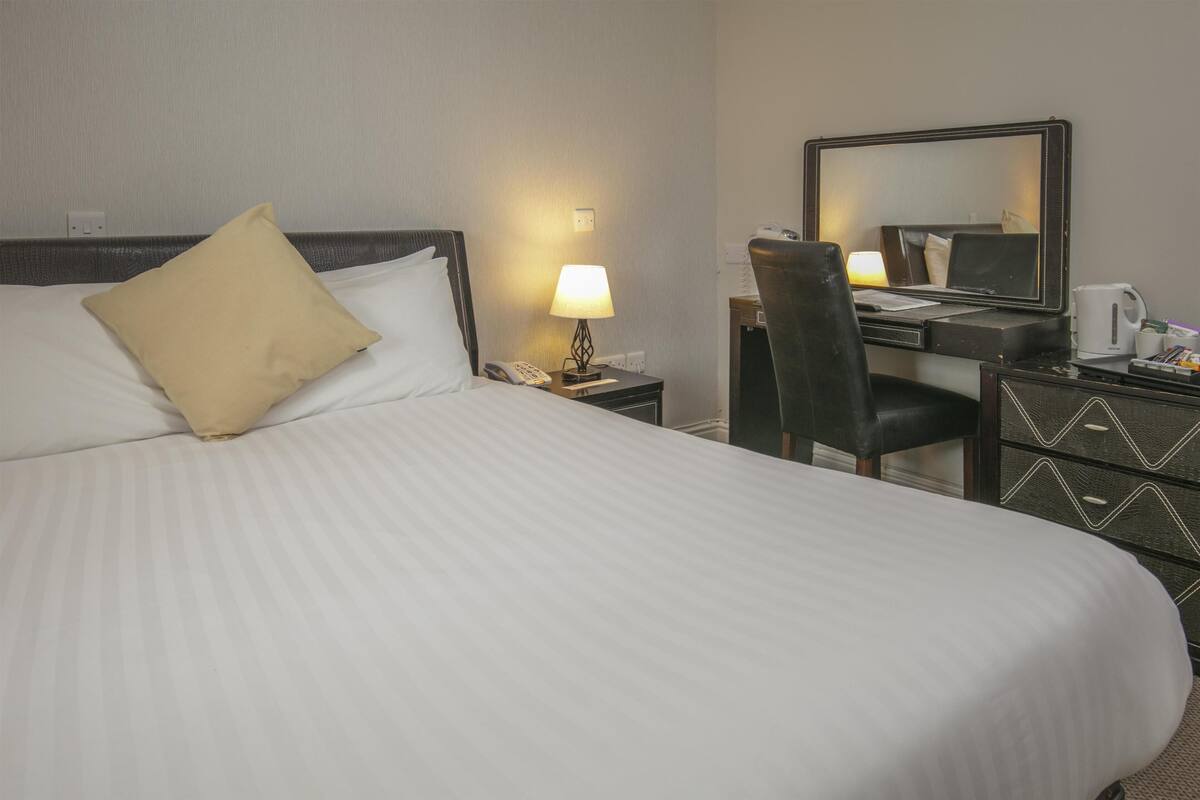 A hotel room with a bed, a desk, a chair, a lamp, a telephone, a mirror, and a coffee maker.