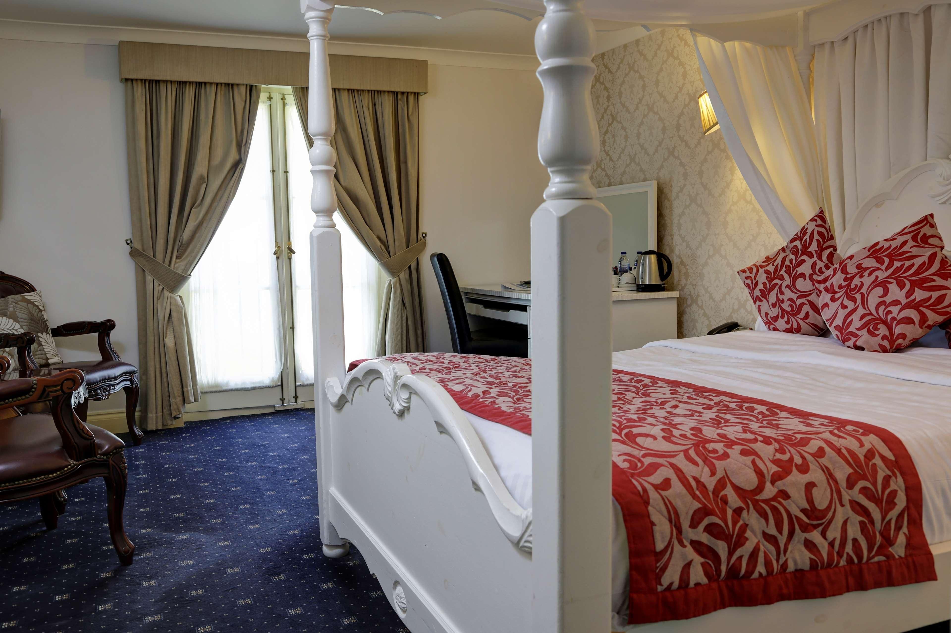 A four-poster bed with a red and white bedspread, a wooden chair, a desk with a lamp, and a window with curtains.
