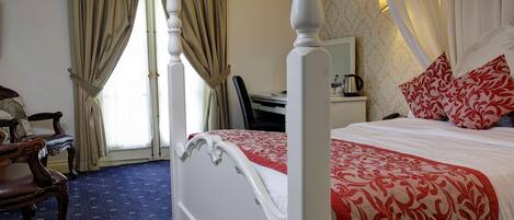 A four-poster bed with a red and white bedspread, a wooden chair, a desk with a lamp, and a window with curtains.