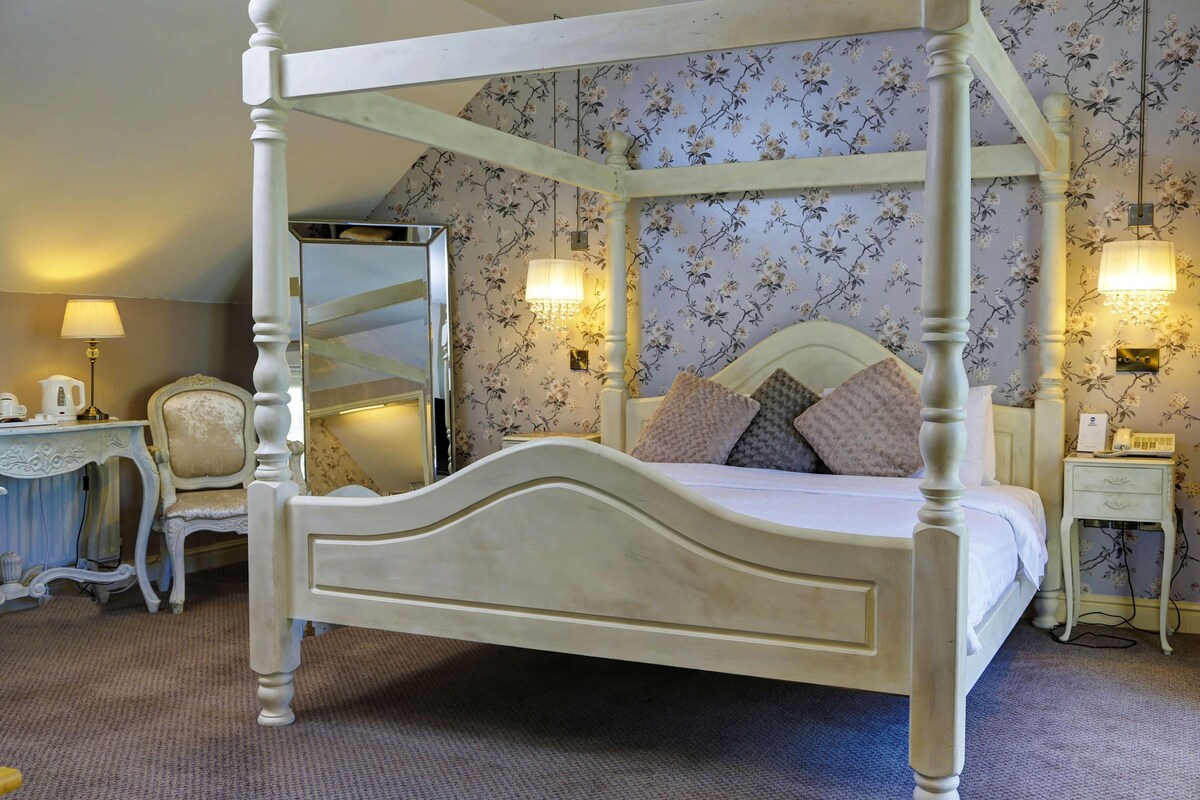 A four-poster bed with a mirror, floral wallpaper, bedside lamps, and a small bedside table with a telephone.