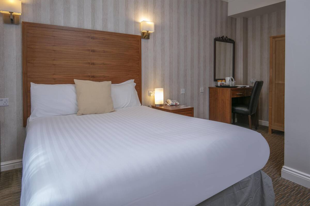 A hotel room with a large bed, a wooden headboard, a desk with a chair, a mirror, and a nightstand with a lamp.