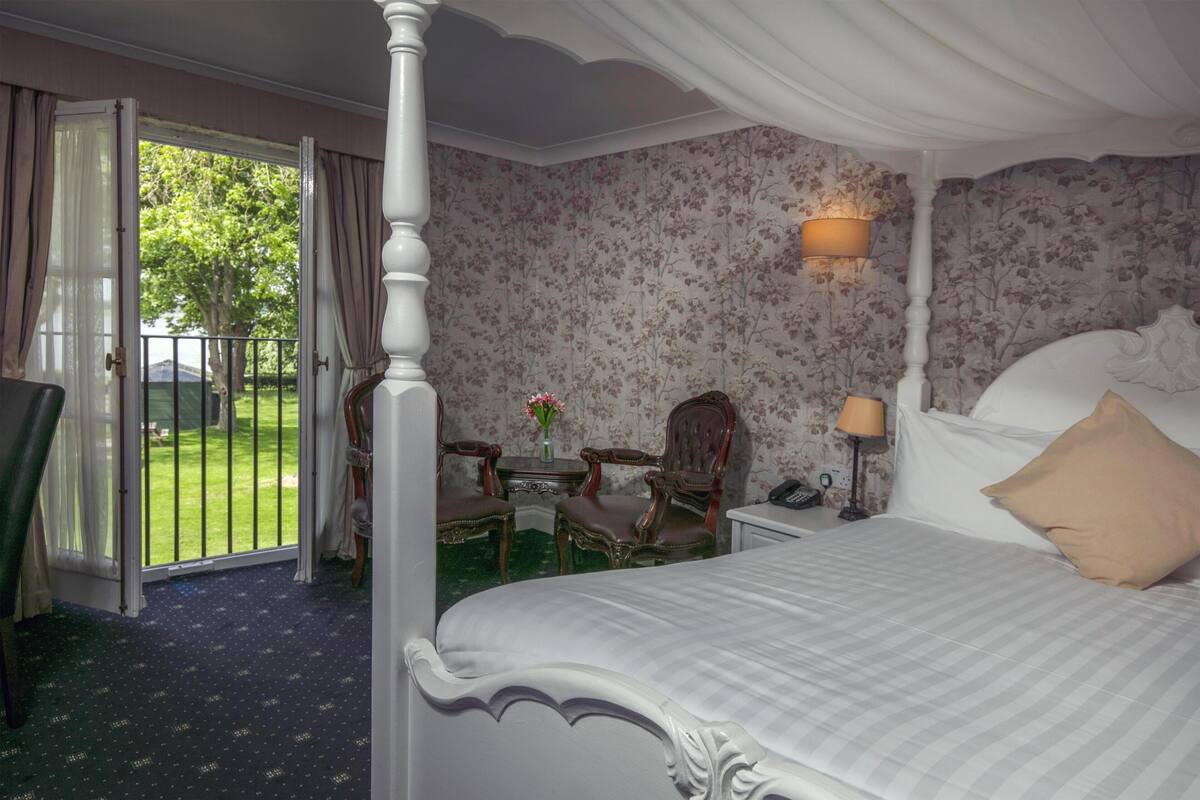 A bedroom with a four-poster bed, a small table with chairs, a lamp, and a view of a green lawn through an open door.