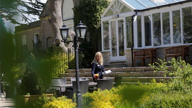 A woman in a black jacket holding a white object, walking past outdoor wicker furniture and a lamp post in a garden area with a conservatory in the background. - Normanton Park Hotel, Sure Hotel Collection by Bes (Oakham)