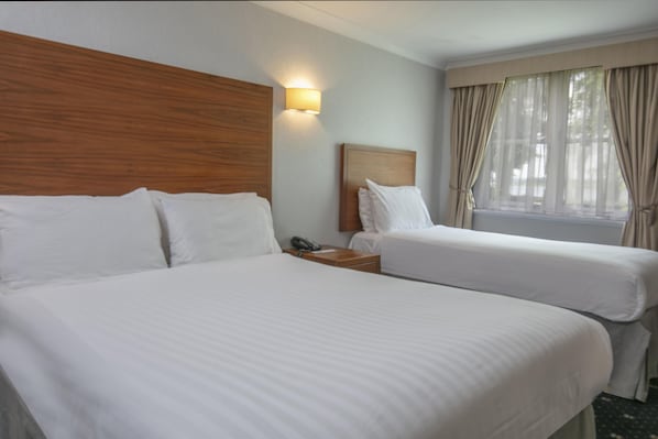 A hotel room with two beds, a wooden headboard, a bedside table with a phone, and a window with curtains.