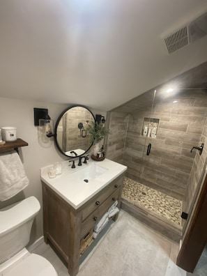 Une salle de bain moderne avec un lavabo blanc, un meuble-lavabo en bois, un miroir rond et une douche séparée par une paroi de verre.
