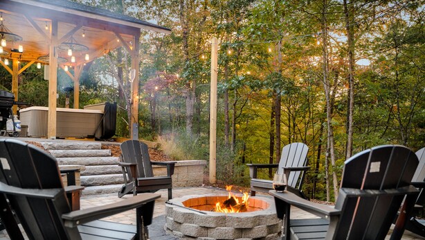 An outdoor patio with Adirondack chairs, a stone fire pit, and a covered seating area with string lights.